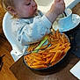 toddler, child, high_chair, pasta, penne, plate, cup, spoon, fork, table, messy, food, eating, sweater, indoor, wood_floor, kitchen, face, person, mealtime