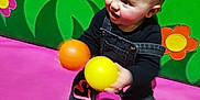 Rae joined the competition — help win amazing prizes! baby, child, denim_overalls, pink_hearts, black_socks, yellow_ball, orange_ball, play_area, green_padded_wall, pink_floor, hand, indoor, cute, happy, playing, toys, infant, smiling, colorful, fun