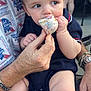 baby, child, toddler, person, hand, face, food, treat, sprinkles, clothing, socks, adult, watch, bracelet, sitting, outdoor, background, blurred_person, festival, feeding