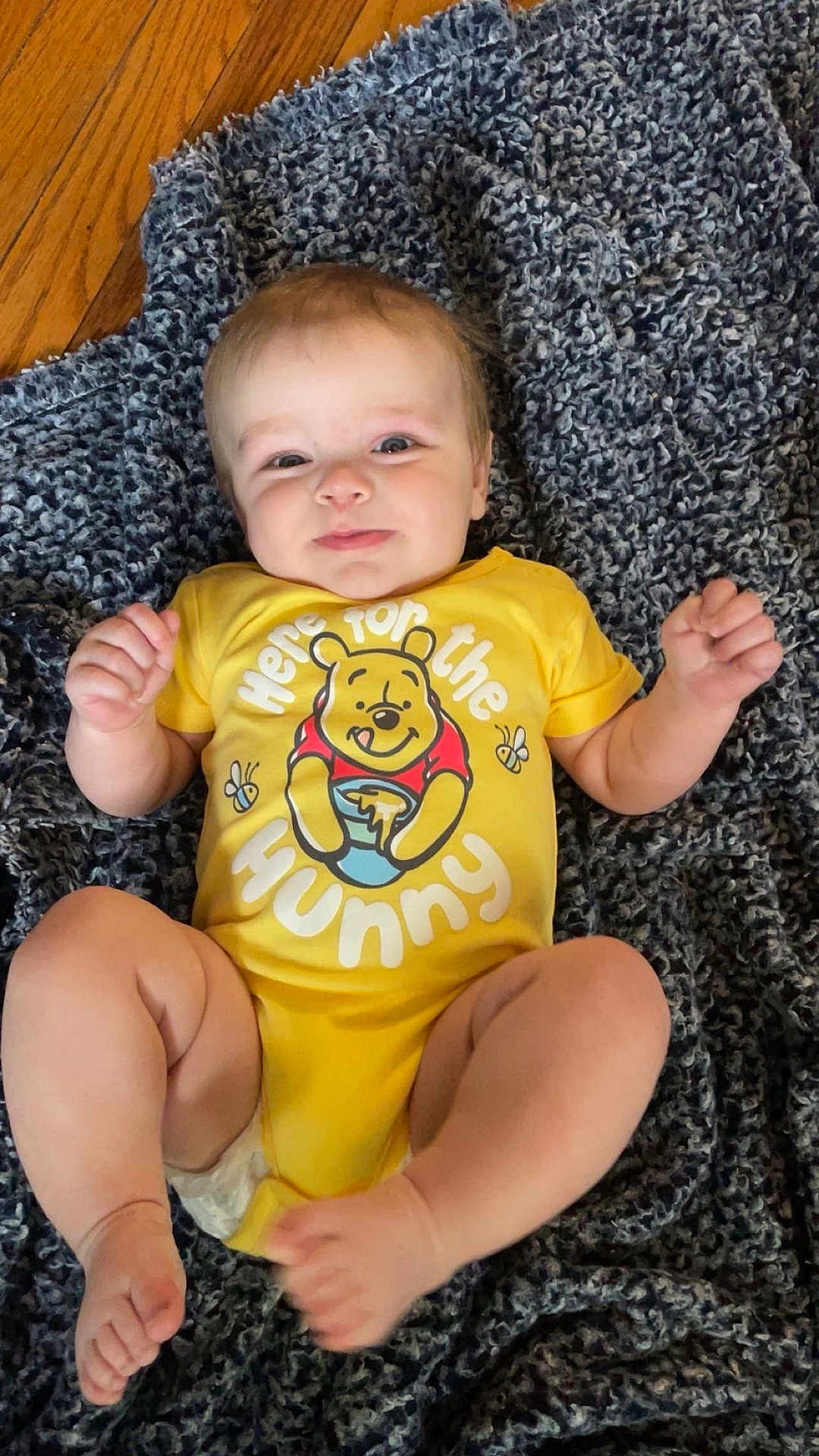 Liam is registered to the contest to win money with this photo: baby, child, infant, yellow_onesie, winnie_the_pooh, blanket, floor, wooden_floor, smile, happy, cute, lying_down, fist, indoors, portrait, toddler, clothing, person, skin, foot
