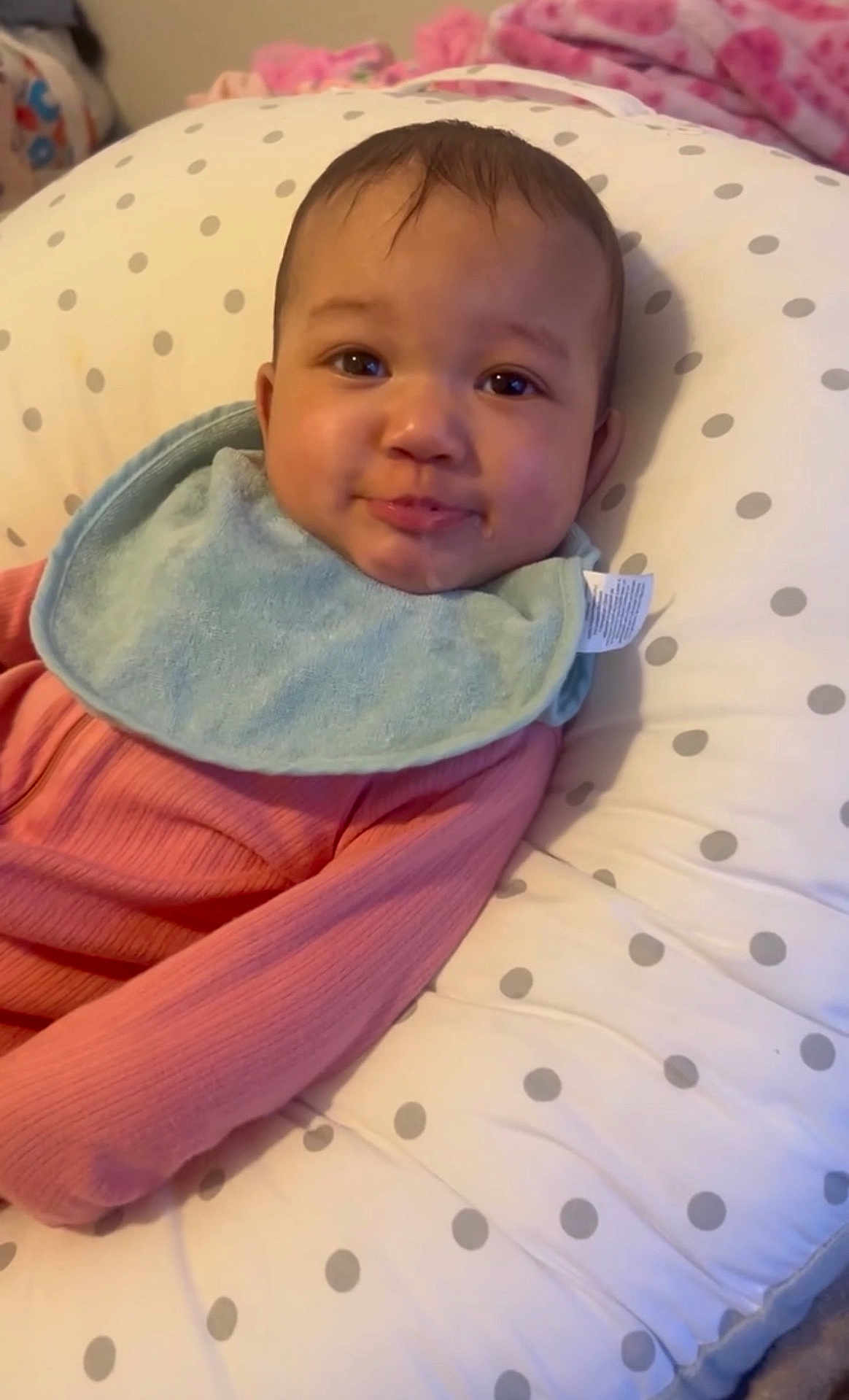 Ameriah is registered to the contest to win money with this photo: baby, infant, bib, pink_clothing, polka_dot, cushion, face, smile, indoors, child, cute, relaxed, soft_fabric, head, young_child, portrait, comfort, baby_bed, sleeping_area, cozy