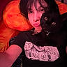 Cheeto is registered to the contest to win money with this photo: person, cat, black_cat, selfie, indoor, orange_background, piercing, tattoo, black_hair, tshirt, pet, necklace, collar, animal, cute, face, eyes, portrait, cozy, closeup