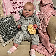 Luella joined the competition — help win amazing prizes! baby, infant, smiling, gray_outfit, pink_blanket, wooden_sign, chair, cozy, cute, happy, indoor, person, child, face, sock, plush_blanket, text_board, baby_milestone, couch, portrait