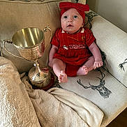 Nyla is registered to the contest to win money with this photo: baby, infant, red_clothing, headband, trophy, couch, blanket, deer_print, indoor, cute, small, sitting, cozy, soft, fabric, face, person, award, wood_floor, home