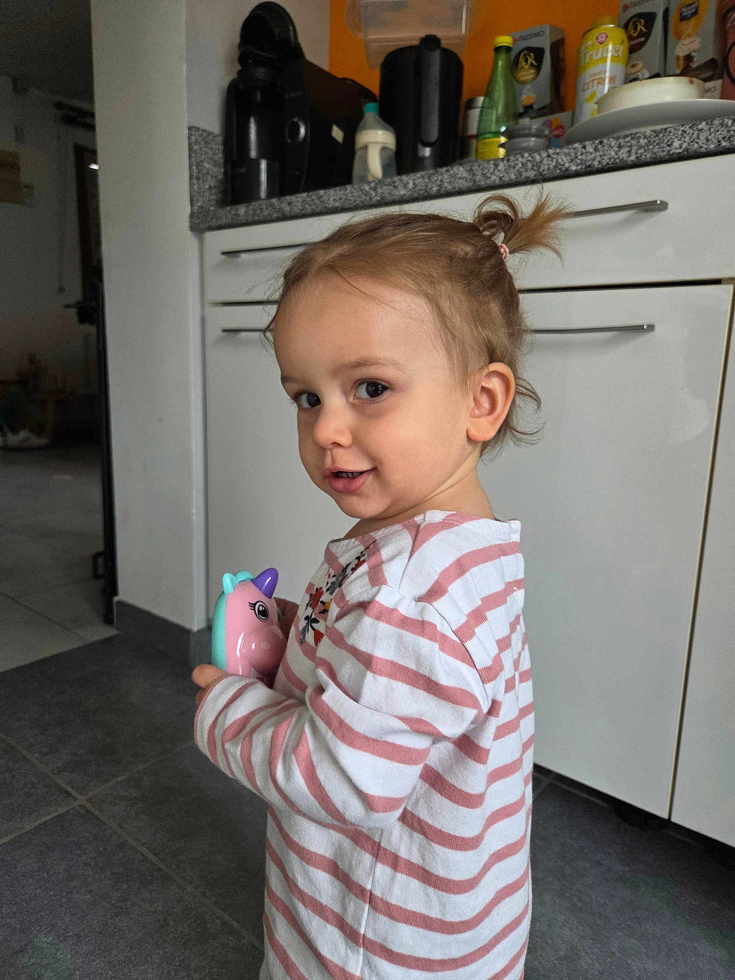 Amalya a rejoint le concours — aidez-le/la à gagner de superbes lots ! child, toddler, toy, unicorn, striped_shirt, pink, white, hair_ponytail, kitchen, cabinet, floor_tile, smile, face, person, indoor, playful, holding, young_child, curious, cute