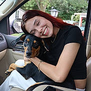 Blicky is registered to the contest to win money with this photo: woman, dog, car_interior, smiling, affection, pet, red_hair, black_dog, tan_markings, casual_clothing, gold_chain, piercing, window, outdoor_background, seat, human_animal_bond, happy, portrait, candid, love