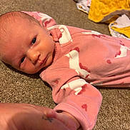 Adeline is registered to the contest to win money with this photo: baby, infant, pink_onesie, carpet, hand_holding, adult_hand, blanket, playpen, floor, person, newborn, skin, face, clothing, indoor, grip, expression, cute, small, child