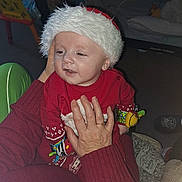Jayden is registered to the contest to win money with this photo: baby, child, santa_hat, red_clothing, adult_hand, indoor, holiday, festive, toy, carpet, sweater, cozy, smiling, cute, infant, person, holding, winter_wear, holiday_season, home