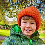 child, smiling, green_jacket, orange_hat, teddy_bear, outdoor, park, trees, grass, autumn, happy, cute, warm_clothing, boy, nature, daylight, portrait, face, childhood, playful