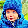 child, blue_hat, knit_hat, puffy_jacket, outdoor, winter_clothing, young_child, face, eyes, smile, curious, warm_clothing, nature_background, portrait, person, cute, happy, headwear, cold_weather, jacket