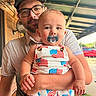 Aryn is registered to the contest to win money with this photo: baby, pacifier, adult, glasses, hat, overalls, american_flag, outdoor, porch, person, child, clothing, facial_hair, smile, hand, arm, window, car, daylight, background
