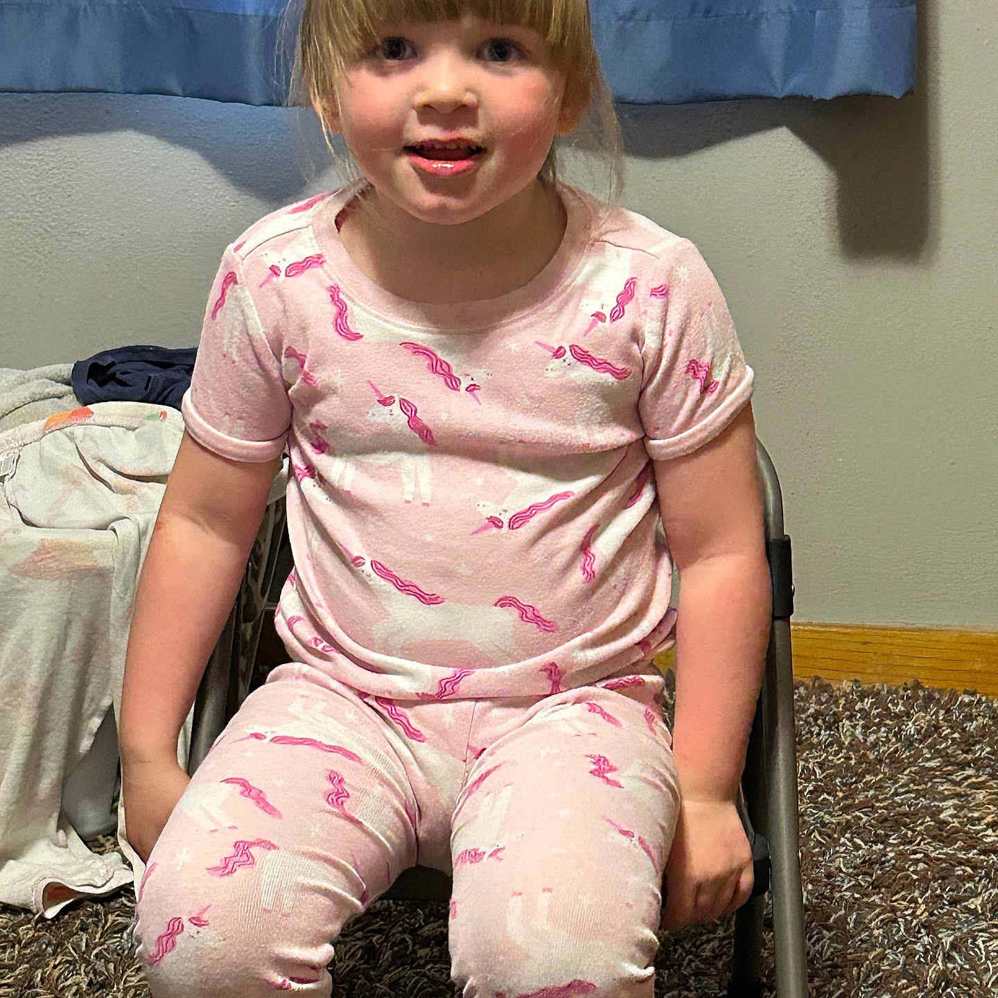 Amber joined the competition — help win amazing prizes! child, pajamas, unicorn, blonde_hair, sitting, chair, carpet, indoor, curtain, smiling, person, young_child, bedroom, casual, home, cute, pale_skin, short_sleeves, pants, relaxed