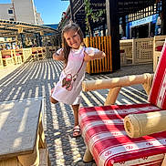 Nicole is registered to the contest to win money with this photo: blue_sky, cafe, chair, child, cobblestone, daylight, girl, happy, outdoor, pattern, person, plant, playful_pose, sandals, shadows, smiling, sunny, table, white_dress, wooden_furniture