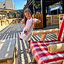 blue_sky, cafe, chair, child, cobblestone, daylight, girl, happy, outdoor, pattern, person, plant, playful_pose, sandals, shadows, smiling, sunny, table, white_dress, wooden_furniture