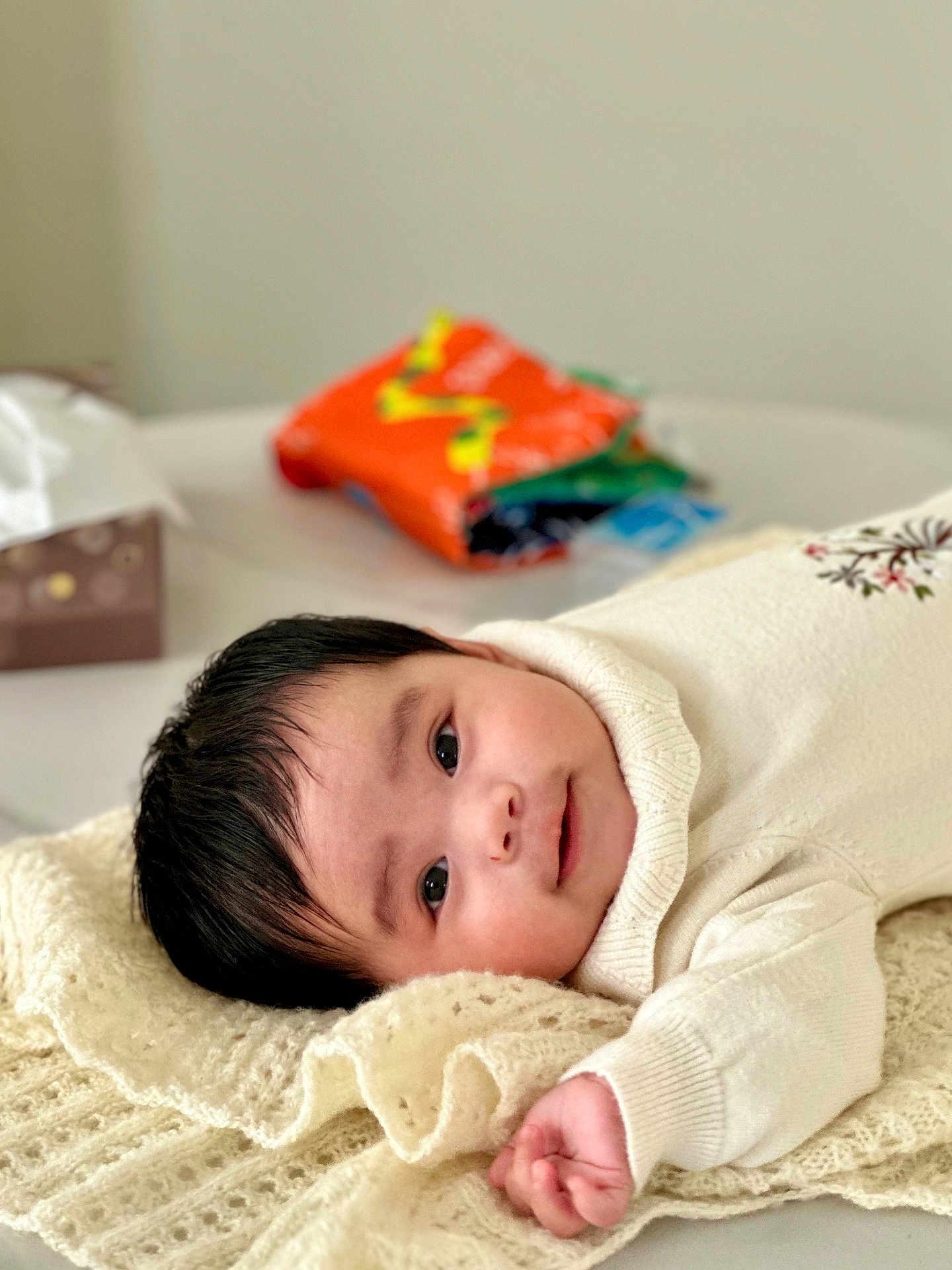 Vivian joined the competition — help win amazing prizes! baby, background_blur, black_hair, blanket, child, curious, cute, face, hand, indoor, infant, knitted, lying_down, portrait, smile, soft_light, table, tissue_box, white_clothing, young