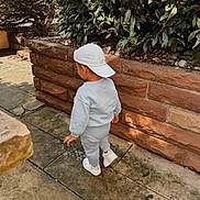 Arilio a rejoint le concours — aidez-le/la à gagner de superbes lots ! toddler, child, back_view, walking, gray_clothing, white_cap, sneakers, stone_path, brick_wall, greenery, bushes, outdoor, sunlight, shadows, casual_wear, sidewalk, nature, daytime, young_child, urban_garden