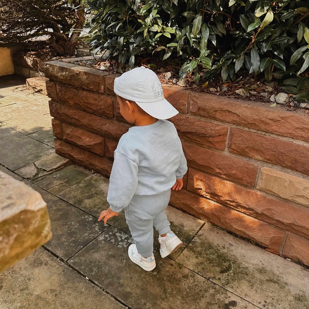 Arilio a rejoint le concours — aidez-le/la à gagner de superbes lots ! back_view, brick_wall, bushes, casual_wear, child, daytime, gray_clothing, greenery, nature, outdoor, shadows, sidewalk, sneakers, stone_path, sunlight, toddler, urban_garden, walking, white_cap, young_child