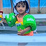 child, toddler, pool, floaties, swimwear, hat, water_toy, curly_hair, outdoor, summer, playing, water, puddle_jumper, green, yellow, blue, palm_trees, background, cute, fun