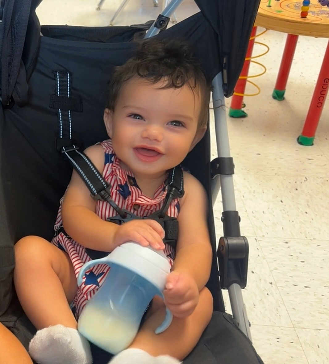 Malakai joined the competition — help win amazing prizes! baby, smiling, stroller, sippy_cup, curly_hair, child, indoor, floor, play_table, toy, clothing, socks, seat_belt, happy, person, infant, face, hands, legs, furniture