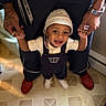 adult, baby, child, floor, footwear, happy, hat, holding_hands, indoor, joyful, overalls, pants, person, portrait, smiling, support, tattoo, toddler, watch, white_shoes