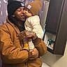 Dreko is registered to the contest to win money with this photo: adult, baby, bathroom, beanie, brown_jacket, cabinet, child, clothing, cozy, facial_expression, holding, home_interior, human, indoor, love, person, portrait, shower_curtain, smiling, toilet