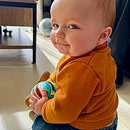 Sandro a rejoint le concours — aidez-le/la à gagner de superbes lots ! baby, ball, cheeks, child, cute, denim_pants, face, floor, furniture, globe, indoor, light, orange_sweater, person, playful, portrait, sitting, smile, toy, window