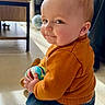 baby, ball, cheeks, child, cute, denim_pants, face, floor, furniture, globe, indoor, light, orange_sweater, person, playful, portrait, sitting, smile, toy, window