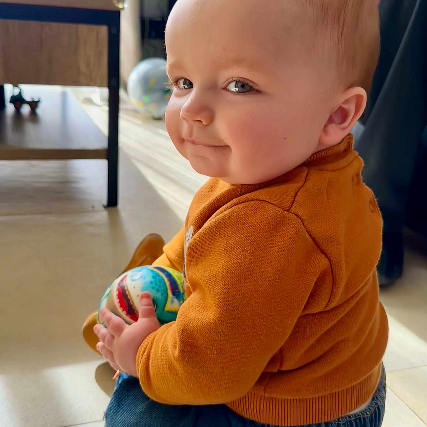 Sandro a rejoint le concours — aidez-le/la à gagner de superbes lots ! baby, ball, cheeks, child, cute, denim_pants, face, floor, furniture, globe, indoor, light, orange_sweater, person, playful, portrait, sitting, smile, toy, window