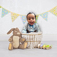 Grayson is registered to the contest to win money with this photo: baby, child, basket, plush_toy, bunny, decorative_eggs, bunting_flags, indoor, floor, white_wall, patterned_shirt, bow_tie, smiling, happy, cozy, soft_blanket, festive, cute, portrait, celebration