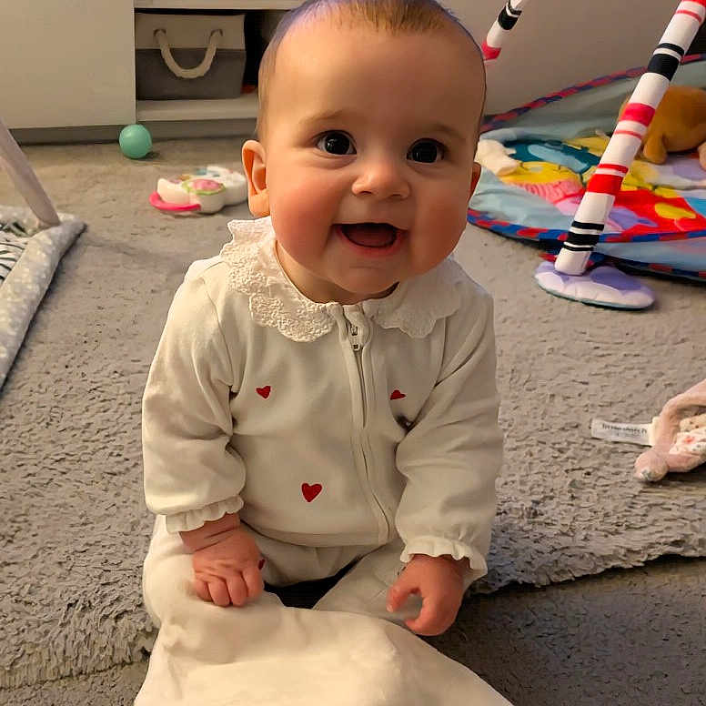 Maisie is registered to the contest to win money with this photo: baby, infant, smiling, white_clothing, heart_pattern, carpet, toys, blanket, playmat, cute, child, indoors, happy, sitting, face, person, soft_lighting, floor, crawling, room
