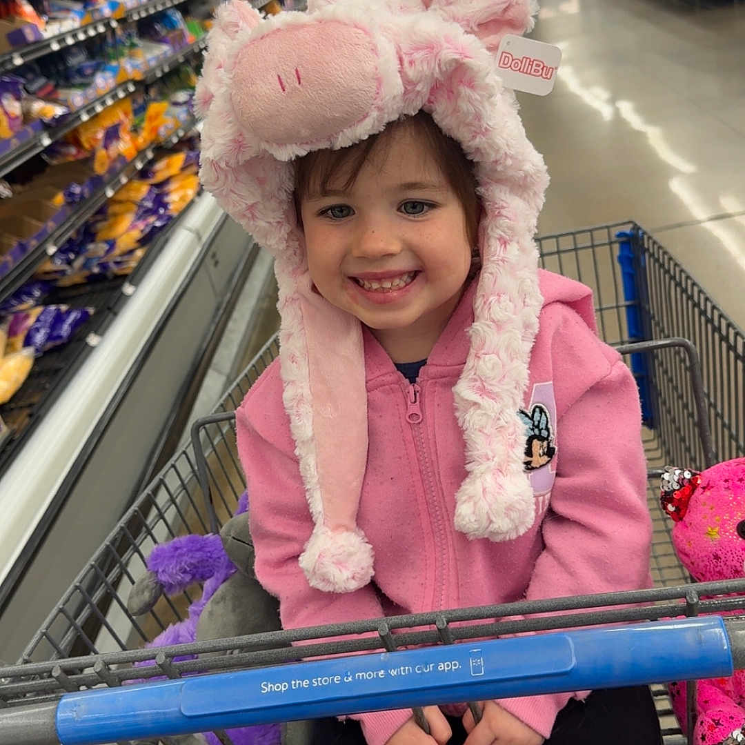 Taytum is registered to the contest to win money with this photo: aisle, casual, child, clothing, cute, floor, grocery_store, happy, hat, indoor, jacket, person, pink_hat, plush_toys, retail, shopping, shopping_cart, smiling, store, toy