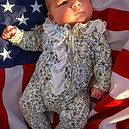 Kimberly is registered to the contest to win money with this photo: baby, infant, american_flag, floral_onesie, sunlight, portrait, cute, child, newborn, soft_light, patterned_clothing, stars, stripes, red, white, blue, lying_down, peaceful, small_hands, adorable