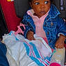 baby, infant, child, car_seat, sitting, denim_jacket, pink_clothing, blanket, pink_cup, wide_eyed, portrait, indoor, vehicle, safety_harness, dark_hair, brown_skin, looking_away, colorful, cozy, photograph