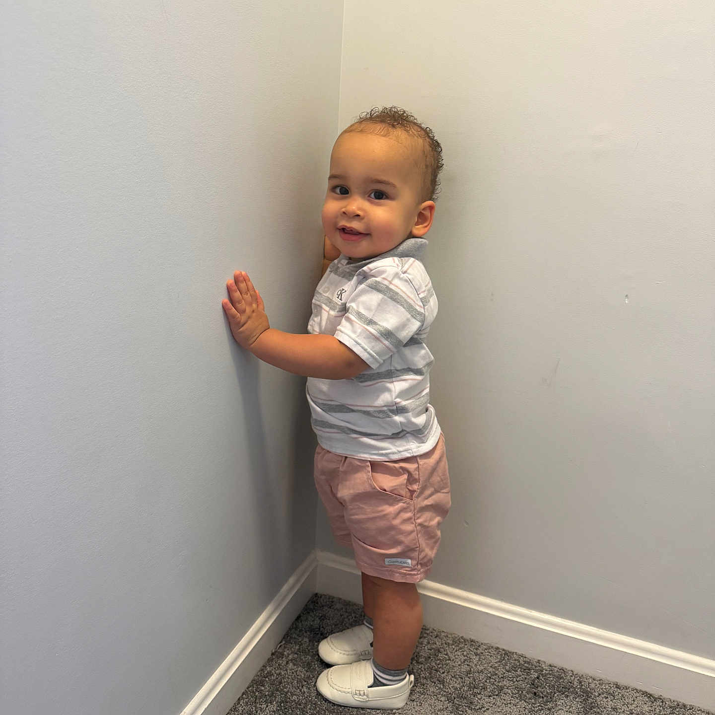 Yaisel is registered to the contest to win money with this photo: toddler, child, boy, standing, corner, wall, carpet, smile, clothing, shirt, shorts, shoes, indoor, curly_hair, cute, happy, person, portrait, casual, light