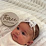 Aria is registered to the contest to win money with this photo: baby, infant, three_months, headband, bow, white_blanket, floral_sweater, face, portrait, lying_down, newborn, cute, soft_texture, milestone, wooden_sign, closeup, child, skin, eyes, peaceful