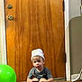 Zayden is registered to the contest to win money with this photo: balloon, casual, child, curious, diaper, door, floor, green_balloon, hat, household, indoor, mat, mop, person, playful, sitting, sock, toddler, toy, wooden_door