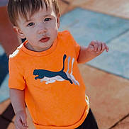 Zayden is registered to the contest to win money with this photo: bright_colors, casual_clothing, cat_graphic, child, curious, cute, daylight, expression, looking_up, orange_shirt, outdoor, pavement, person, portrait, short_hair, small_child, standing, sunlight, toddler, young_child
