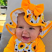 Lily-Mae is registered to the contest to win money with this photo: baby, child, sunflower, hat, bow, yellow_clothing, smiling, happy, outdoor, cushion, face, portrait, infant, sitting, nature, bright_colors, cute, young_child, headwear, joyful