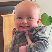 Ivy is registered to the contest to win money with this photo: adult_hand, baby, cheeks, close_up, ear, eyes, face, hands, happy, indoor, infant, onesie, pajamas, pink_nails, plant, portrait, skin, smiling, wooden_furniture, zipper