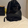 cat, black_and_white_cat, pet, animal, backpack, bag, bed, bedroom, indoors, sitting, fluffy, whiskers, feline, portrait, travel, luggage, blanket, white_bedding, door, cozy