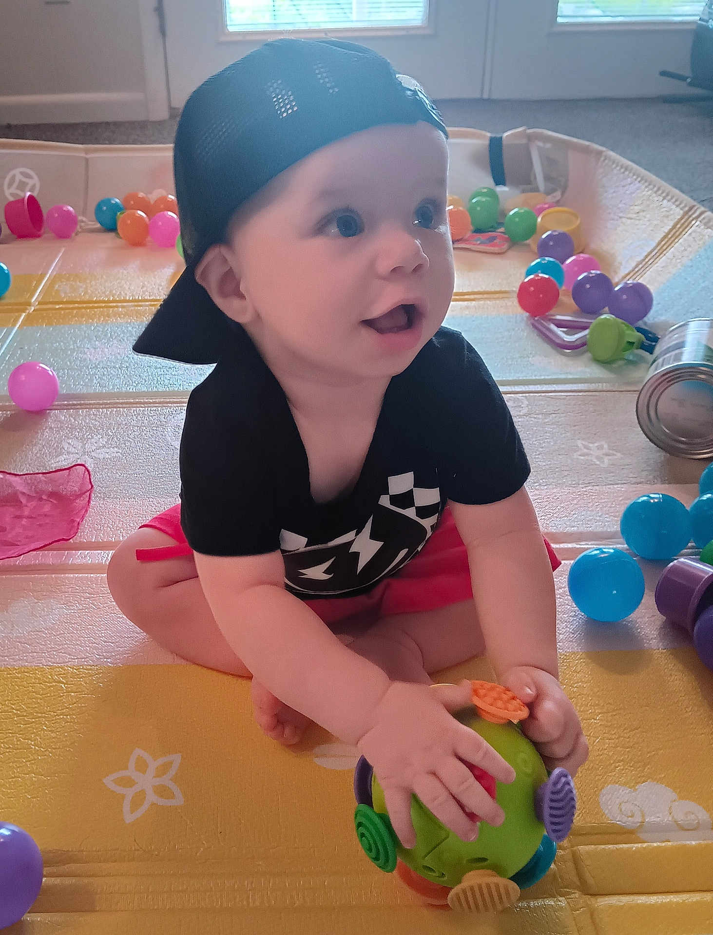 Slade joined the competition — help win amazing prizes! baby, child, cap, toy, play_mat, plastic_balls, indoor, cute, happy, sitting, colorful, clothing, hand, face, mouth_open, eyes, floor, room, window, light