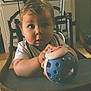 baby, child, high_chair, toy, ball, face, person, wooden_chair, indoor, furniture, table, photograph, cabinet, curious, expression, holding, playtime, room, home, seated