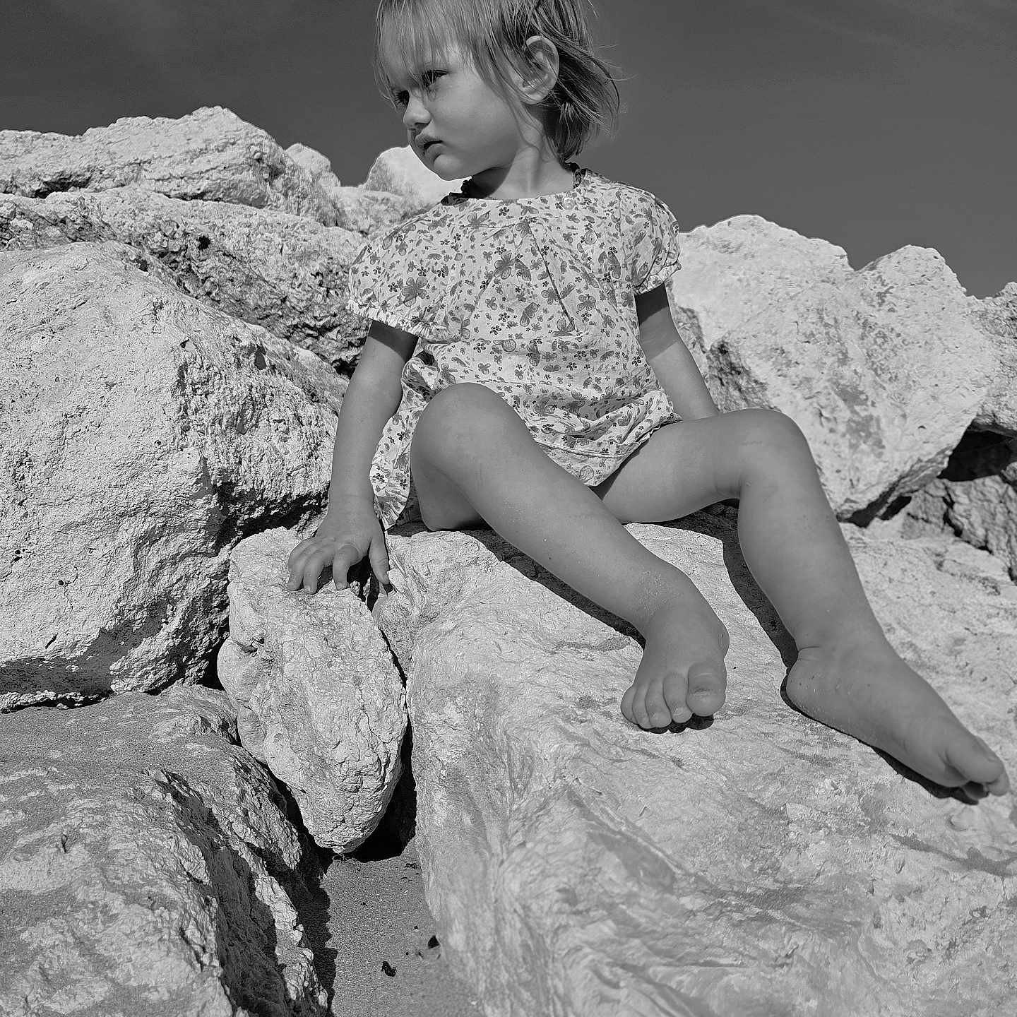 Lou a rejoint le concours — aidez-le/la à gagner de superbes lots ! bare_legs, barefoot, casual, child, daylight, dress, monochrome, nature, outdoor, portrait, rocks, sand, serious_expression, side_view, sitting, sky, small_person, sunlight, toddler, young_child