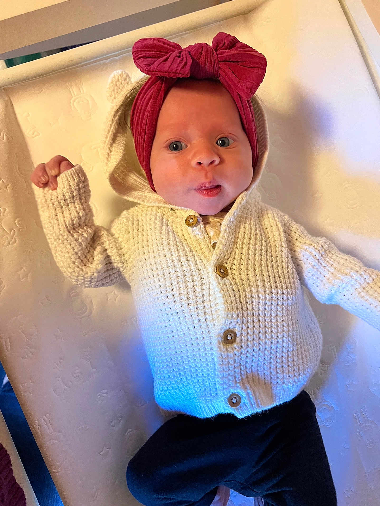 Sadie is registered to the contest to win money with this photo: baby, infant, knitted_sweater, headband, red_bow, white, black_pants, lying_down, curious_expression, closeup, indoor, soft_lighting, cute, child, portrait, face, newborn, texture, warm_clothing, cozy