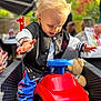 toddler, child, toy_car, red, blue, black_shoes, jacket, outdoor, daylight, happy, blond_hair, smiling, steering_wheel, furniture, teddy_bear, garden, colorful, people, celebration, party