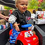 Lyzio participe au concours pour gagner de l'argent avec cette photo : toddler, child, toy_car, spiderman, smiling, outdoor, party, jacket, pants, black_shoes, curly_hair, red, blue, plastic_toy, happy, playful, gift_bags, blurred_background, person, seat