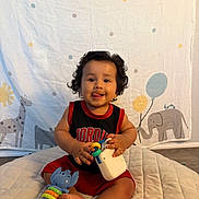 Emmanuel joined the competition — help win amazing prizes! baby, child, curly_hair, smiling, tongue_out, sitting, toy, blue_toy, red_clothing, black_clothing, quilted_mat, giraffe, elephant, balloons, background, indoor, cute, playful, happy, portrait