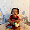 baby, child, curly_hair, smiling, tongue_out, sitting, toy, blue_toy, red_clothing, black_clothing, quilted_mat, giraffe, elephant, balloons, background, indoor, cute, playful, happy, portrait