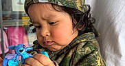 Emmanuel joined the competition — help win amazing prizes! toddler, child, toy, blue_toy, camouflage_clothing, hoodie, cap, curly_hair, indoor, person, hand, face, focused, cute, casual_clothing, play, closeup, expression, holding, background