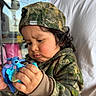 toddler, child, toy, blue_toy, camouflage_clothing, hoodie, cap, curly_hair, indoor, person, hand, face, focused, cute, casual_clothing, play, closeup, expression, holding, background